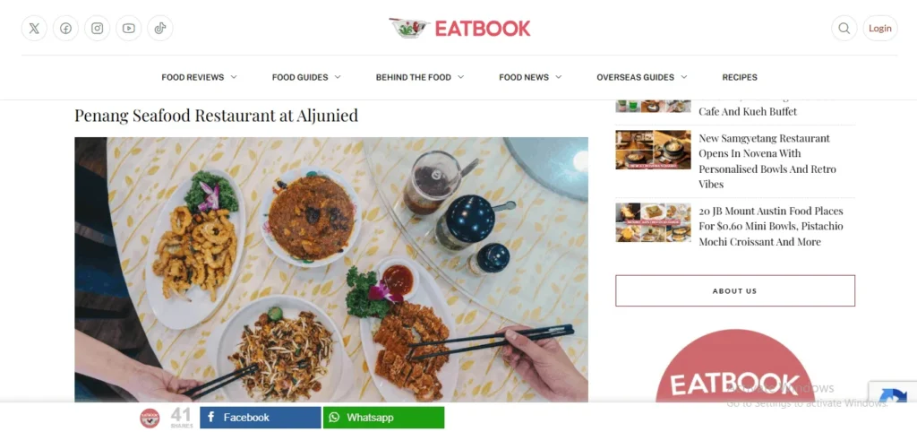 Screenshot of the Eatbook website featuring an article titled "Penang Seafood Restaurant at Aljunied." The featured photograph shows an overhead view of a family-style meal spread on a yellow floral tablecloth, including fried calamari, stir-fried noodles, a curry dish, and crispy pork rolls, with two hands holding chopsticks reaching toward the plates.