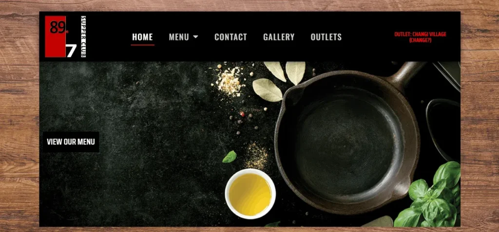 Website homepage for "89.7 Supper Club".
The top navigation bar is black and includes a logo in a red box with the white numbers 89 and 7. The menu options are in white text and include "Home", "Menu", "Contact", "Gallery", and "Outlets". There is also red text in the top-right that reads "Outlet: Changi Village (Change?)".
The background is a dark, rough texture, with various spices, including pepper and dried bay leaves, scattered around the top. A black cast iron pan sits on the right. In the bottom-left corner is a white cup filled with oil and next to it some basil leaves. There is also white text on the left in a small black box that reads "View our Menu".