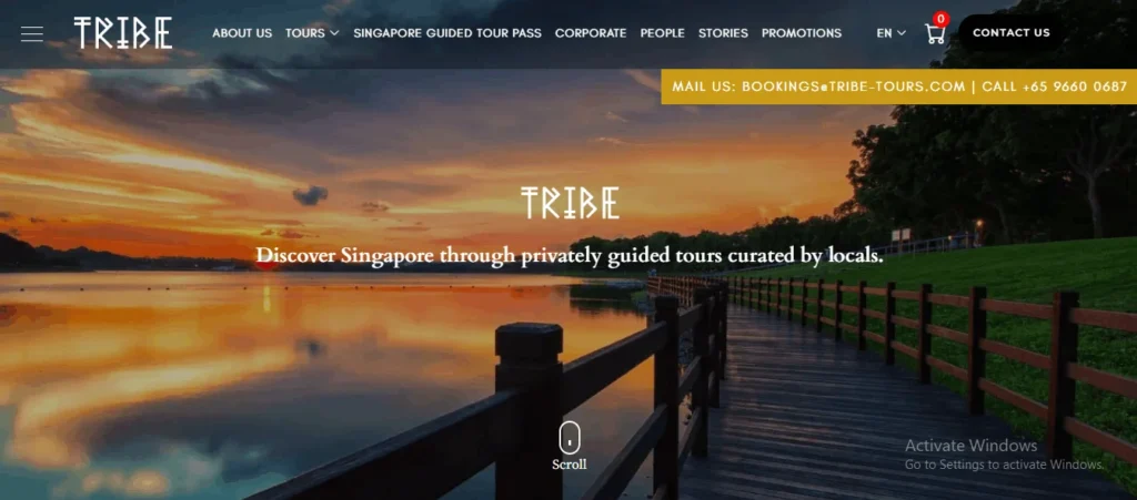 Homepage for TRIBE featuring a scenic background image of a Singapore sunset over a park lake with a wooden boardwalk. Large white text overlays read "TRIBE. Discover Singapore through privately guided tours curated by locals." and a top navigation bar with a yellow contact banner is visible. The logo TRIBE is in the top left. A scroll icon is at the bottom center.