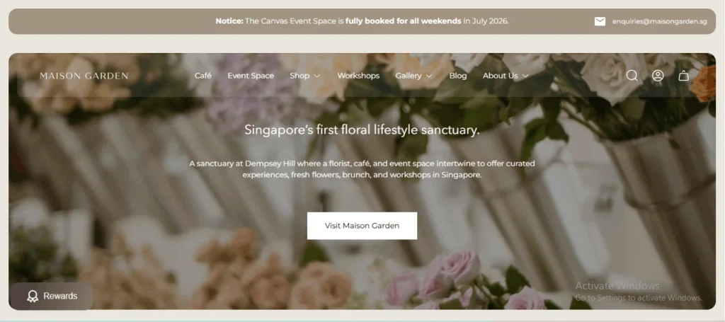 Website homepage header for Maison Garden, featuring a soft-focus background of pastel flowers, navigation menu links, and a central tagline describing it as "Singapore’s first floral lifestyle sanctuary."
