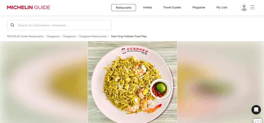 Michelin Guide website layout showing top navigation options and a search bar, with breadcrumbs leading to "Nam Sing Hokkien Fried Mee". The main banner highlights a top-down view of fried Hokkien noodles served with prawns, egg, and a small dish of cut red chili and lime on a pink plate.