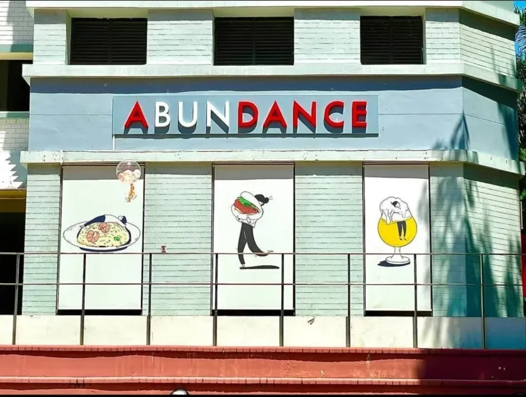 Exterior storefront of a restaurant named "ABUNDANCE" in bold, 3D red capital letters on a pastel light-blue brick building. Below the sign, three large window panels display whimsical, minimalist illustrations: on the left, a person relaxes on a giant plate of fried rice; in the center, a person carries a massive gua bao (steamed pork belly bun); and on the right, a person is draped lazily over the foam of a giant glass of beer.