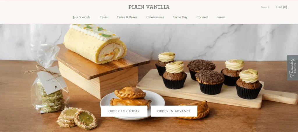 Plain Vanilla Bakery homepage banner showcasing a spread of artisanal bakes on wooden boards: a cream-filled roll cake, chocolate and frosted cupcakes, pastries, and green kaya cookies in a clear gift bag. Includes navigation menu items (July Specials, Cafés, Cakes & Bakes, Celebrations) and buttons for "Order For Today" and "Order In Advance."