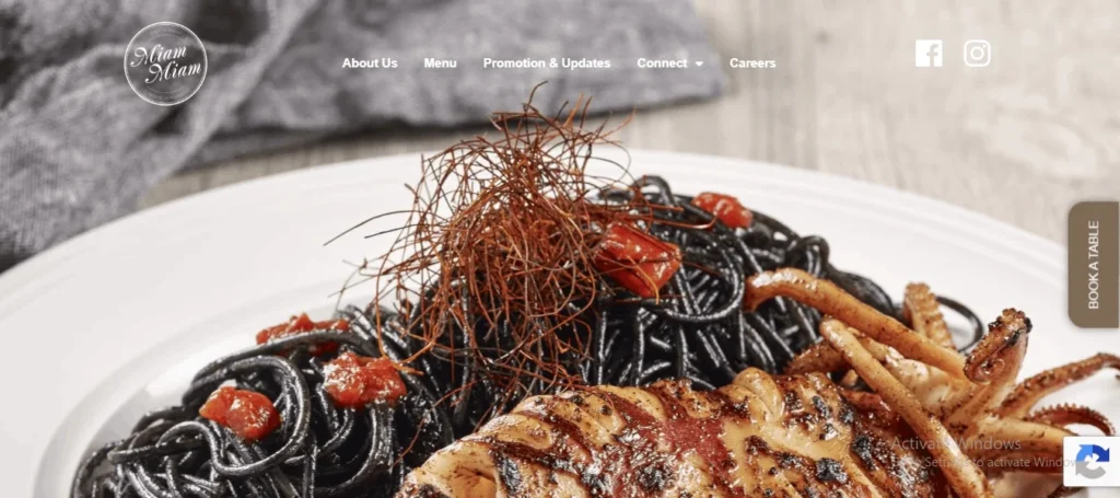 Website banner for Miam Miam French-Japanese restaurant at Westgate Singapore, showcasing a signature dish of black squid ink pasta topped with fine chili threads and served with a whole grilled squid.