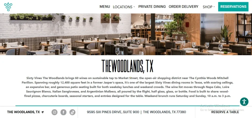 A website screenshot for Sixty Vines in The Woodlands, TX, featuring a top navigation menu and a teal "Reservations" button. Below the menu is a wide banner image showing a modern, upscale restaurant interior with light wooden tables set for dining, a white brick wall with small potted wall plants, and an open kitchen in the background. Beneath the photo, a stylized header reads "THE WOODLANDS, TX" followed by descriptive text about the restaurant's wine tap selection, space, and menu. The bottom section includes social media icons, the restaurant's address, and a "RESERVE A TABLE" link.
