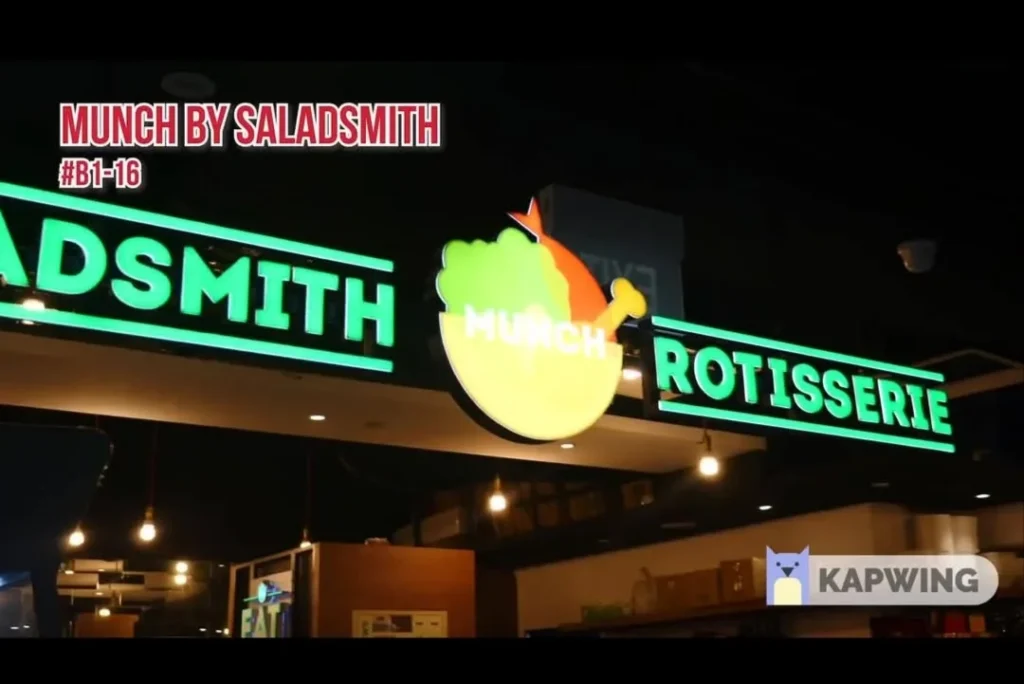 Illuminated green and yellow storefront sign for Munch Saladsmith Rotisserie unit #B1-16 inside a shopping mall.